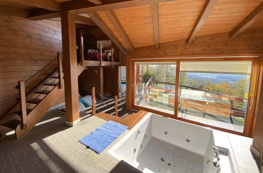 Foto dell'interno della struttura dello Chalet Relax Dolce Benessere, terzo centro benessere in ordine di costruzione del Resort Spa La Bossola di Netro. In primo piano la vasca idromassaggio, a sinistra una scala in legno che porta nel soppalco attrezzato come zona relax con angolo bar. Di fronte alla vasca idromassaggio una doppia porta a vetri a scorrimento in legno che offre una vista sul paesaggio boschivo della serra morenica di Ivrea.