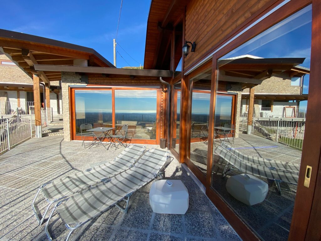 Nella fotografia, in primo piano lo Chalet Dolce Benessere, il terzo centro benessere in ordine di edificazione del Resort Spa La Bossola con la sua struttura in legno e le sue ampie vetrate che riflettono il paesaggio della serra morenica di Ivrea, il cielo terso ed azzurro ed un ampio terrazzo con un tavolino e due sedie per godersi un aperitivo e due sdraio per prendere il sole.