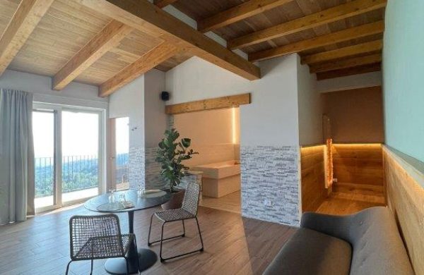 Vista dall'interno del centro benessere Magici Momenti, quarto chalet relax di recente costruzione del Resort Spa La Bossola. il salotto si presenta con tetto e pavimento in legno; le pareti sino a quasi metà altezza sono decorate con pietre a vista e legno. In fondo alla stanza si vede la stanza del sale e la vetrata scorrevole che porta sul balcone privato con vista sulla serra morenica di Ivrea e sul Monviso.