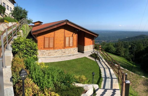 Nella fotografia in primo piano si vede lo Chalet Vacanza Lauretana, circondata da aiuole e piantine, si arriva all'ingresso percorrendo una scalinata in pietra con corrimano in legno. La struttura è in legno con un muretto decorativo alla sua base, di fronte allo chalet un prato con erba rasata. In secondo piano una vista sul paesaggio verde della serra morenica di Ivrea e di Mongrando.